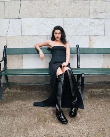 Woman in a black strapless dress and tall leather boots seated on a green park bench against a stone wall.