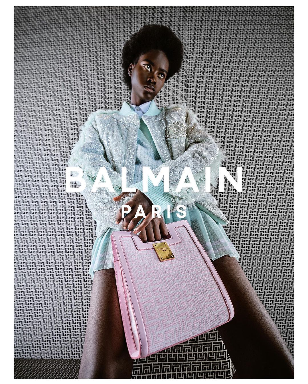 Fashion shot featuring a Black model wearing a sequined jacket, mini skirt, and carrying a pink handbag against a patterned backdrop.