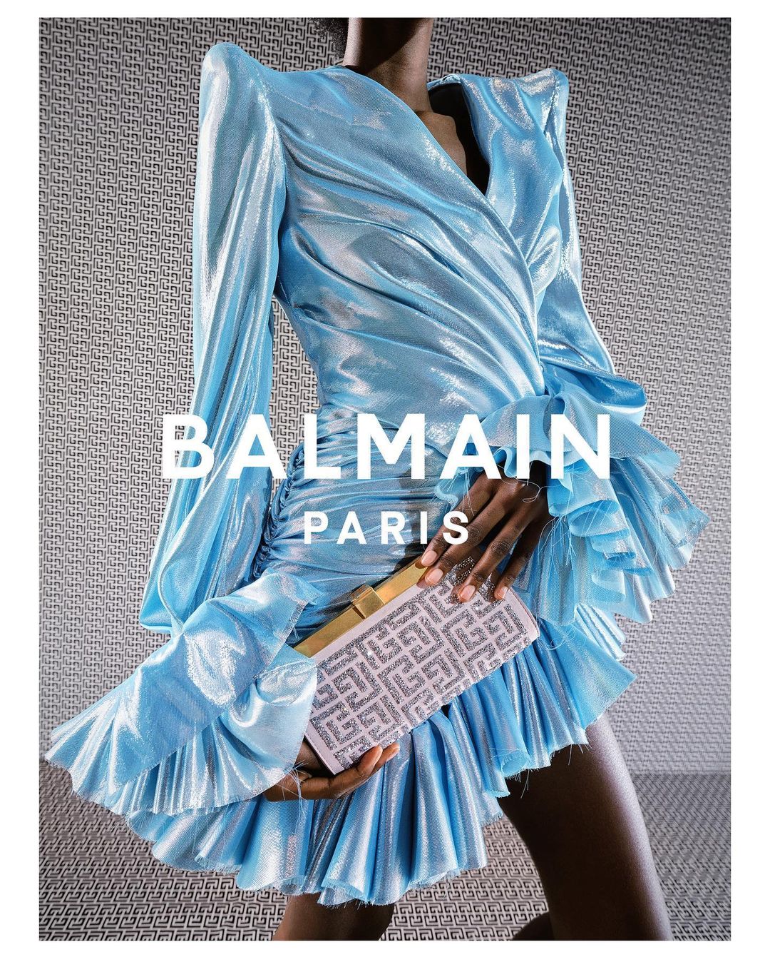 A woman in a light blue Balmain dress holds a clutch, showcased against a textured gray backdrop. The dress features long sleeves and ruffles.