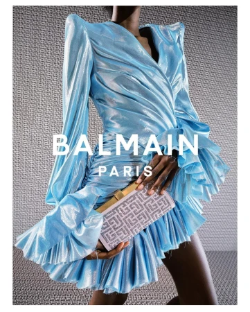A woman in a light blue Balmain dress holds a clutch, showcased against a textured gray backdrop. The dress features long sleeves and ruffles.
