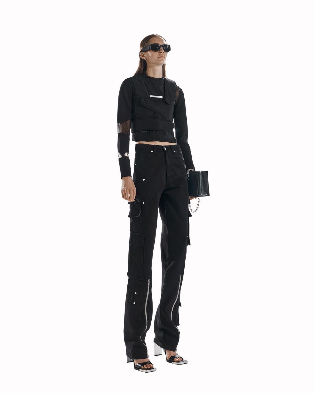 Fashionable woman poses in black top and cargo pants with silver details, holding a matching black purse against a white backdrop.