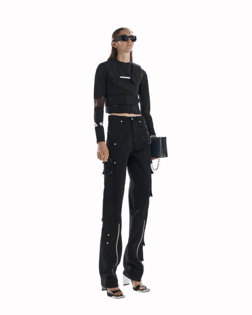 Fashionable woman poses in black top and cargo pants with silver details, holding a matching black purse against a white backdrop.