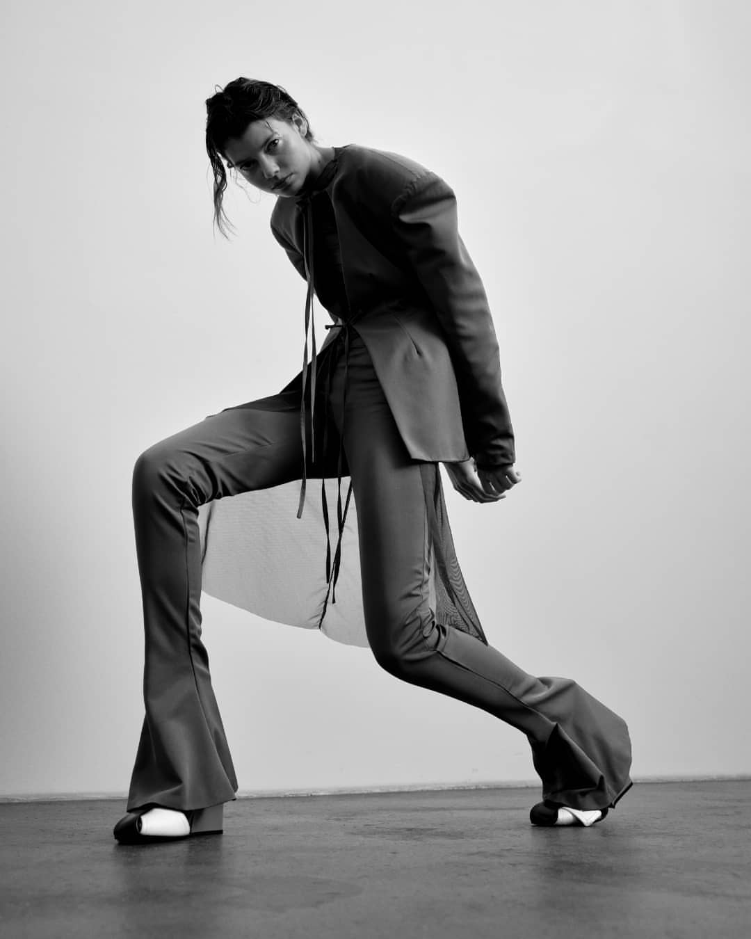 Monochrome fashion shot of a woman in an oversized blazer and flared pants, leaning back against a light gray wall in a studio setting.