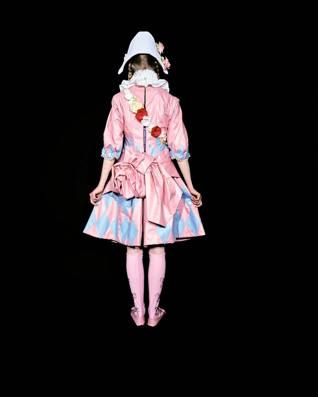 Back view of person wearing a pink and blue diamond-patterned dress, matching socks and shoes, and a white cap against black backdrop.