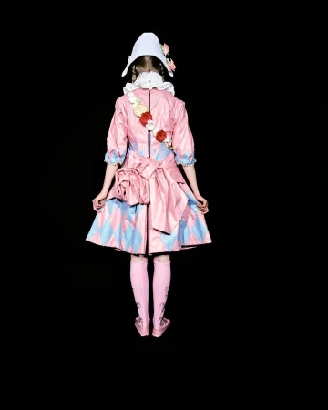 Back view of person wearing a pink and blue diamond-patterned dress, matching socks and shoes, and a white cap against black backdrop.