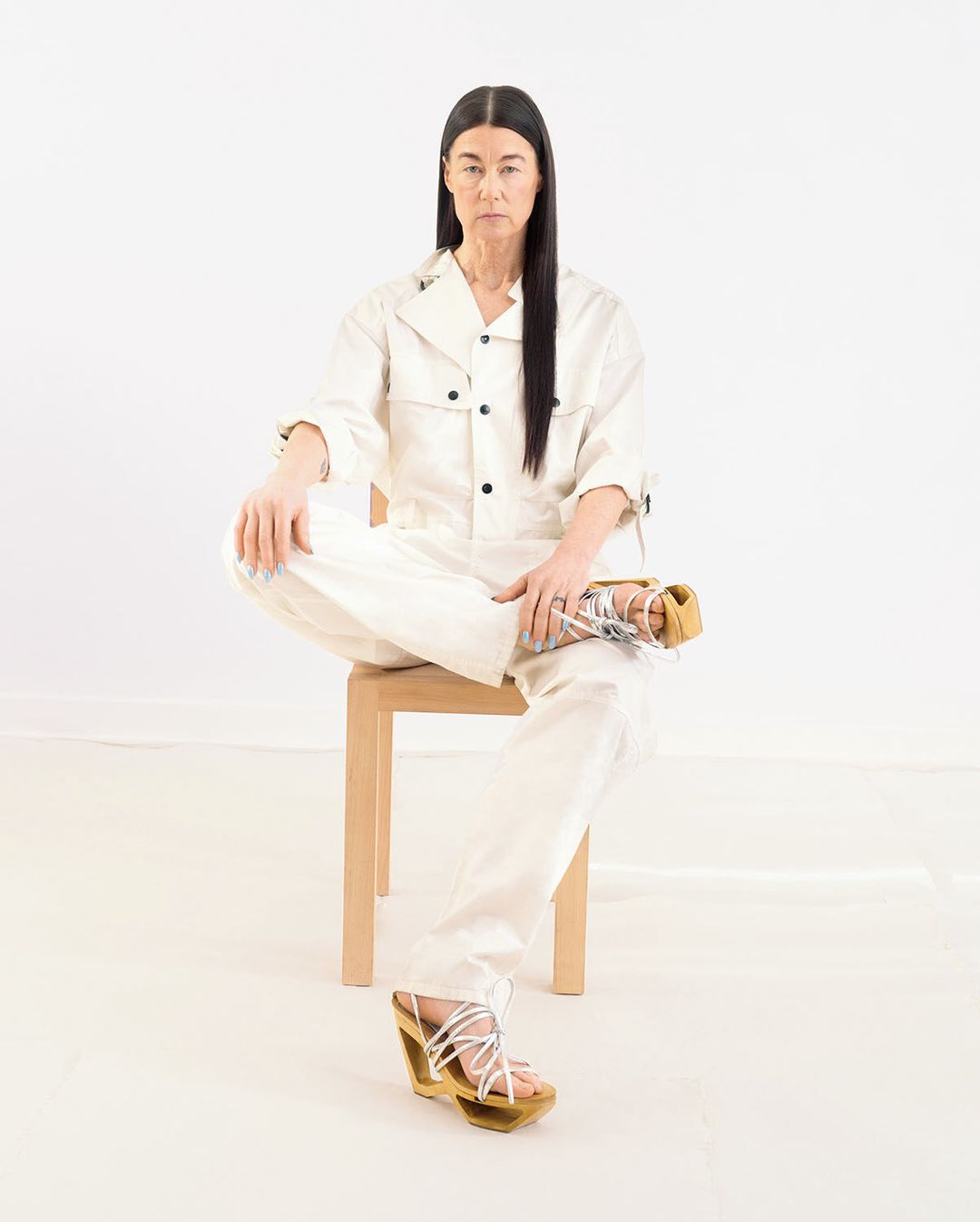 A woman in a light beige jumpsuit sits cross-legged on a chair against a white background, wearing wedge sandals with tie-up straps.