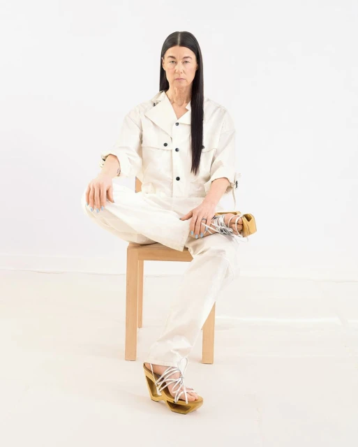 A woman in a light beige jumpsuit sits cross-legged on a chair against a white background, wearing wedge sandals with tie-up straps.