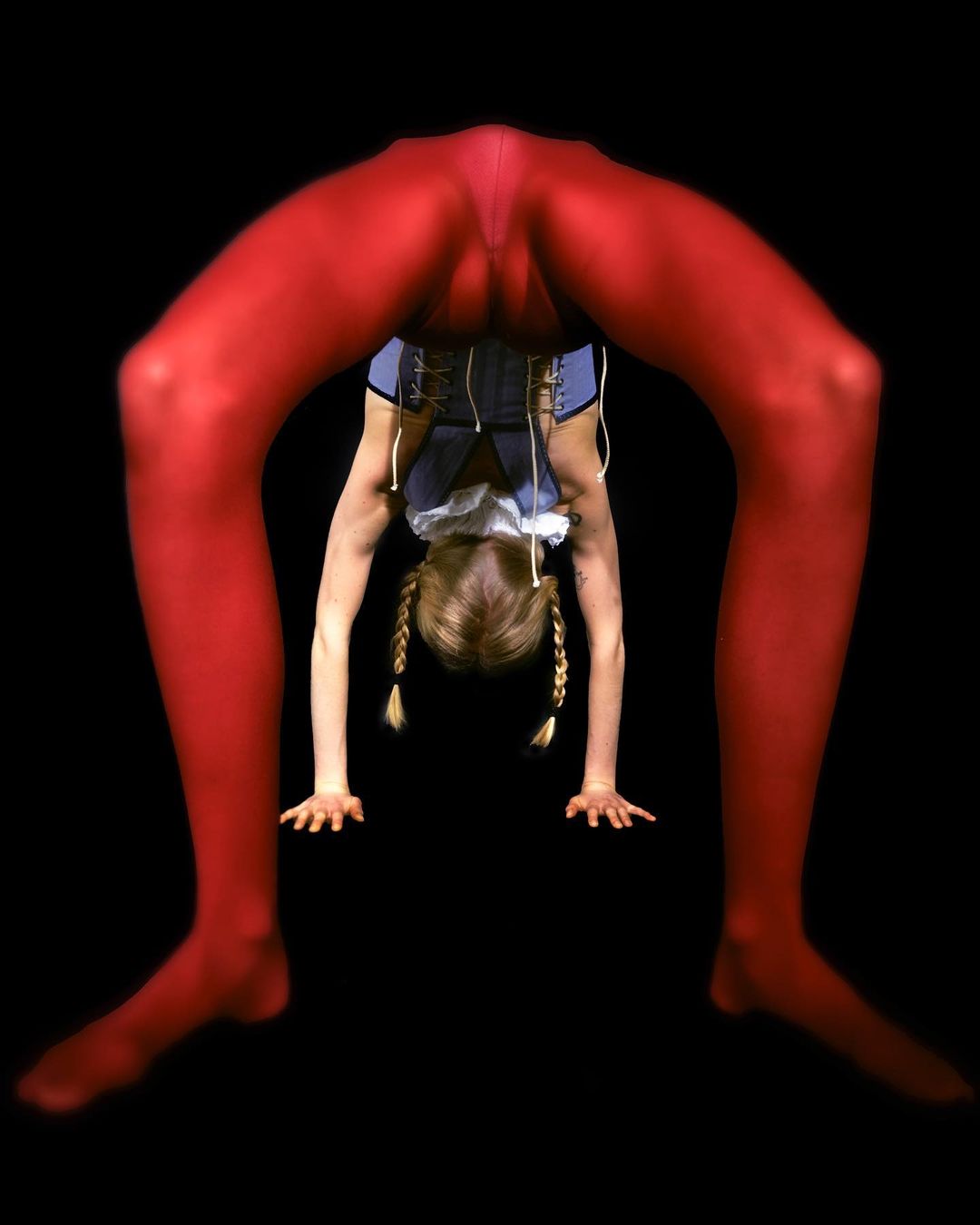 Young female gymnast in red leggings performing a backbend, viewed through her legs, against a stark black backdrop in a studio setting.
