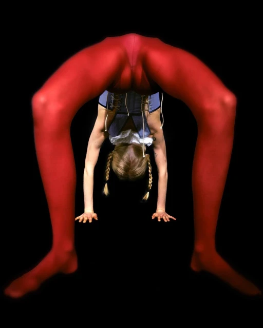 Young female gymnast in red leggings performing a backbend, viewed through her legs, against a stark black backdrop in a studio setting.