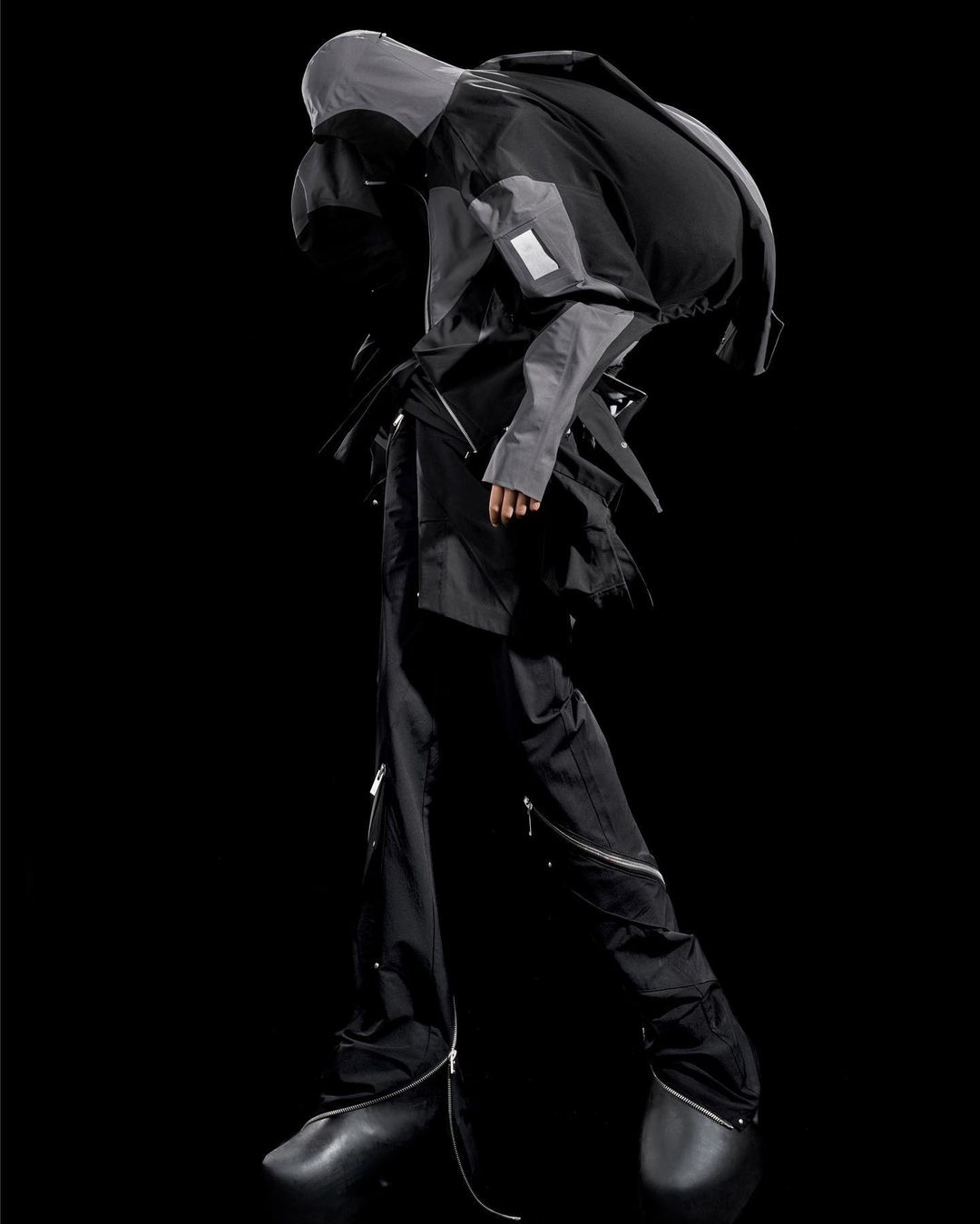 Fashion model showcases avant-garde grey hooded jacket and black pants with zippers in a studio shot, featuring unique black shoes.
