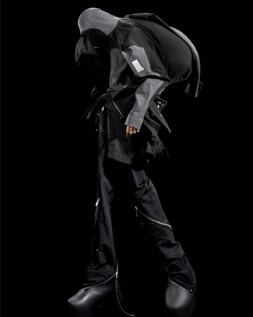 Fashion model showcases avant-garde grey hooded jacket and black pants with zippers in a studio shot, featuring unique black shoes.