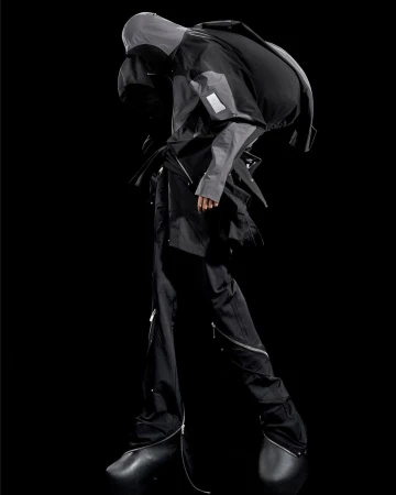 Fashion model showcases avant-garde grey hooded jacket and black pants with zippers in a studio shot, featuring unique black shoes.