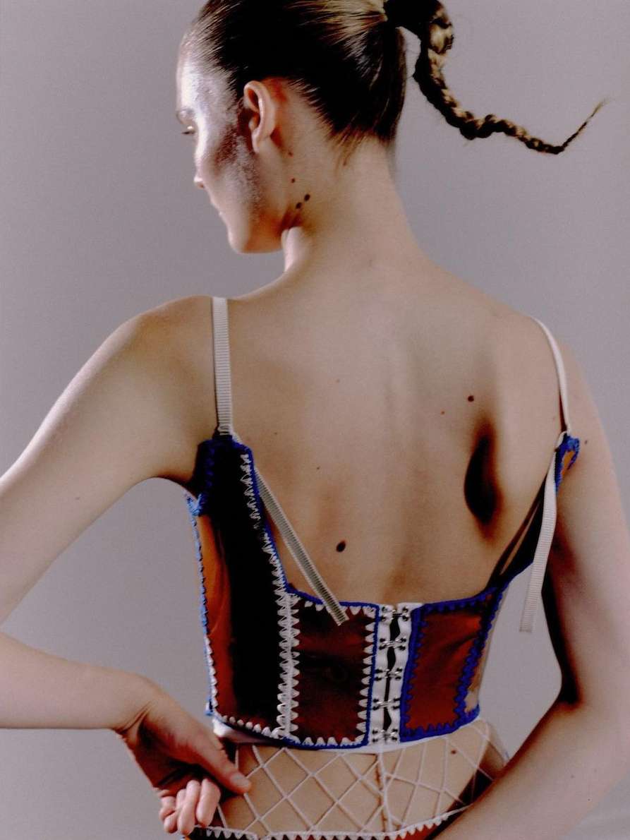 Back view of a fashion model wearing a brown and blue corset with a white skirt, showcasing braided hair against a plain background.