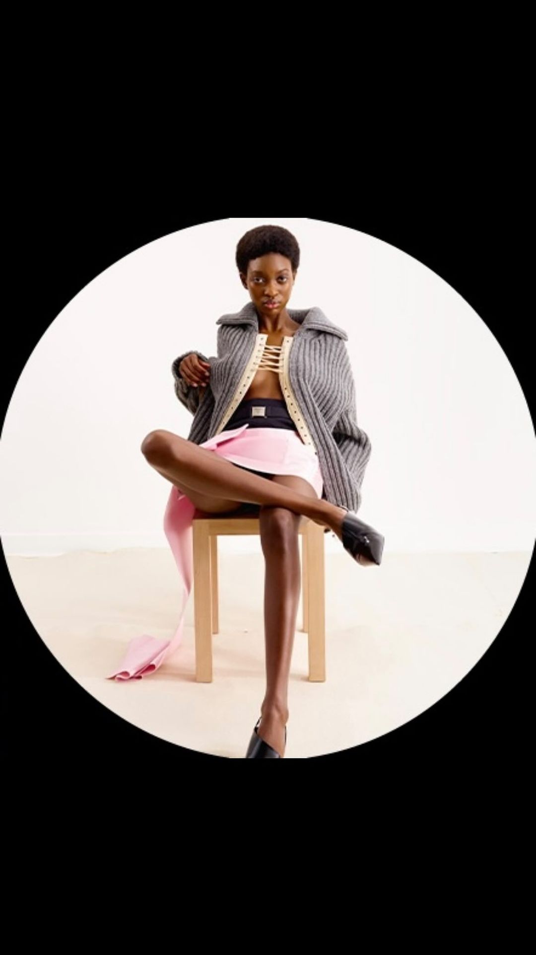 A dark-skinned woman with short hair is sitting on a stool wearing a grey coat, black top, and pink skirt in a studio setting.