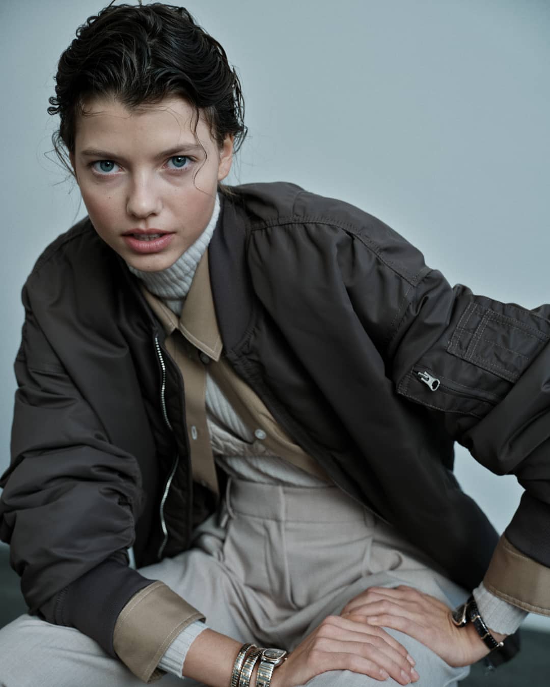 Portrait of a young woman with wet dark hair wearing a bomber jacket, turtleneck, and bracelets, looking directly at the camera.