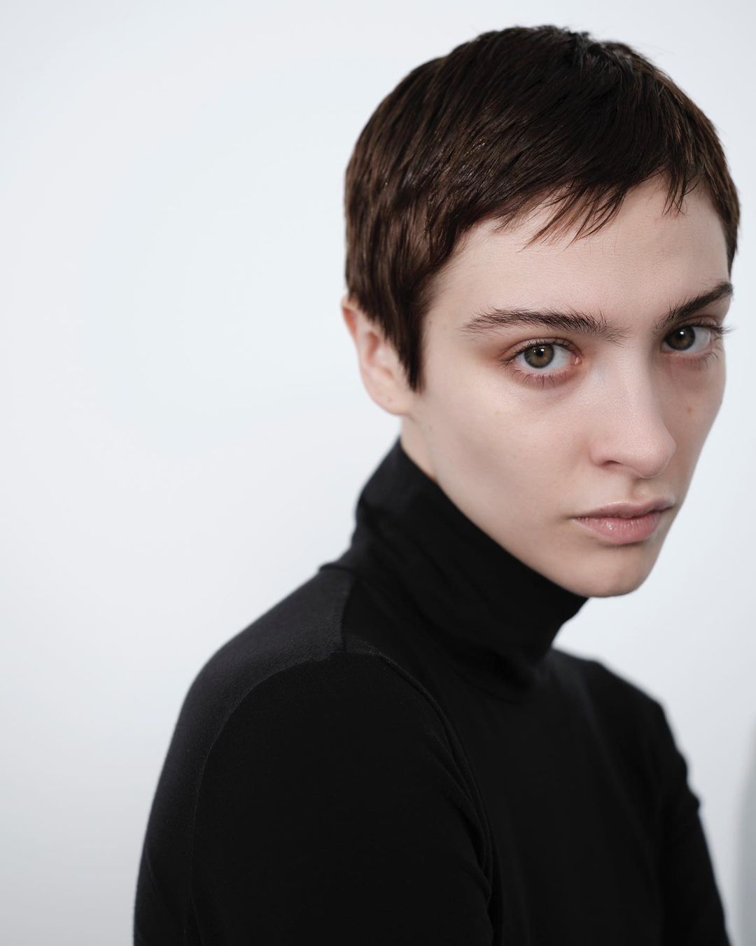 Close-up portrait of a young adult woman with short brown hair wearing a black turtleneck sweater against a white background.
