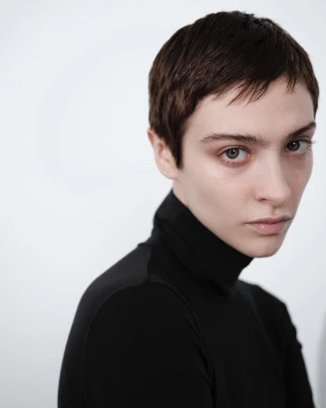 Close-up portrait of a young adult woman with short brown hair wearing a black turtleneck sweater against a white background.