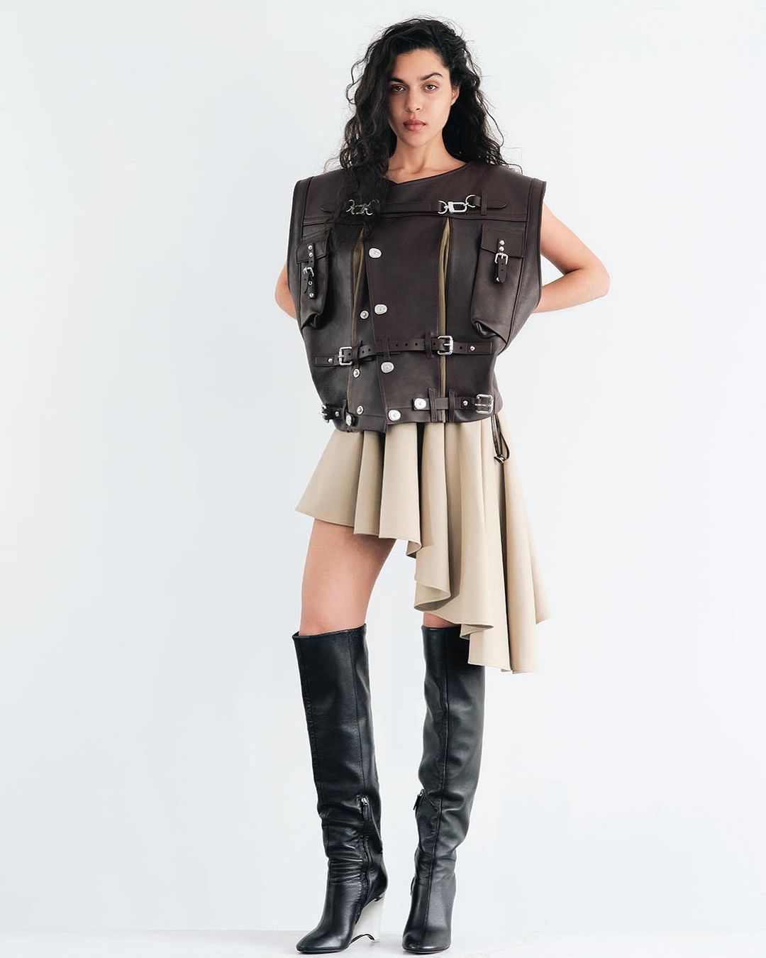 A woman poses in a brown leather vest, beige skirt, and black boots against a white backdrop in a fashion-forward studio portrait.