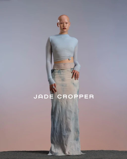Bald woman in a light blue top and long tie-dye skirt poses in a studio with gradient background and textured flooring. Fashion editorial.