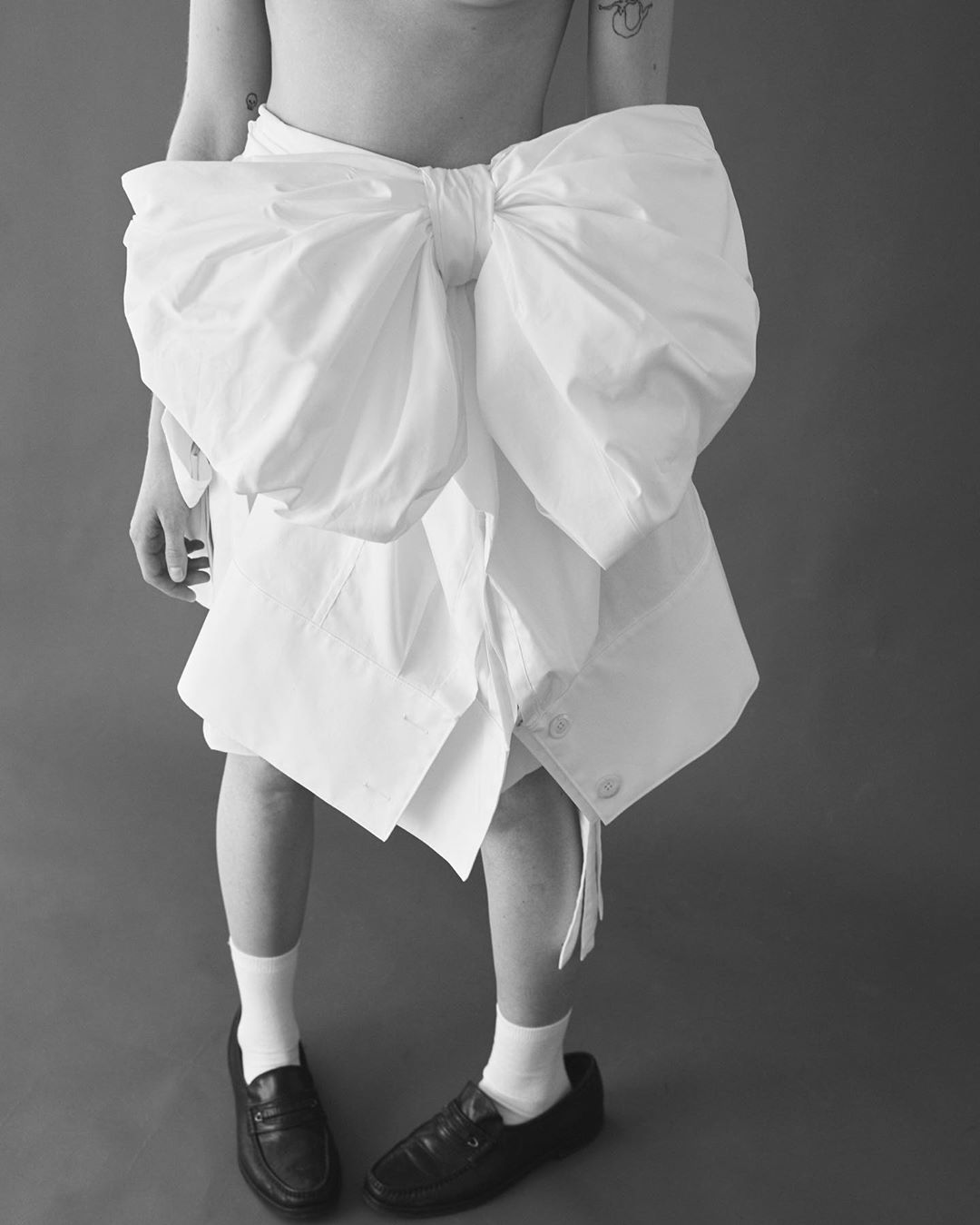 Studio shot of an adult wearing an oversized white bow and partial skirt with black shoes against a grey backdrop in black and white.