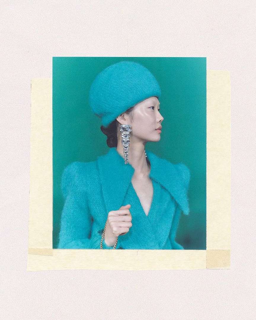 Woman in a teal textured coat and matching beret poses in a studio with a bright green background, accessorized with statement earrings.
