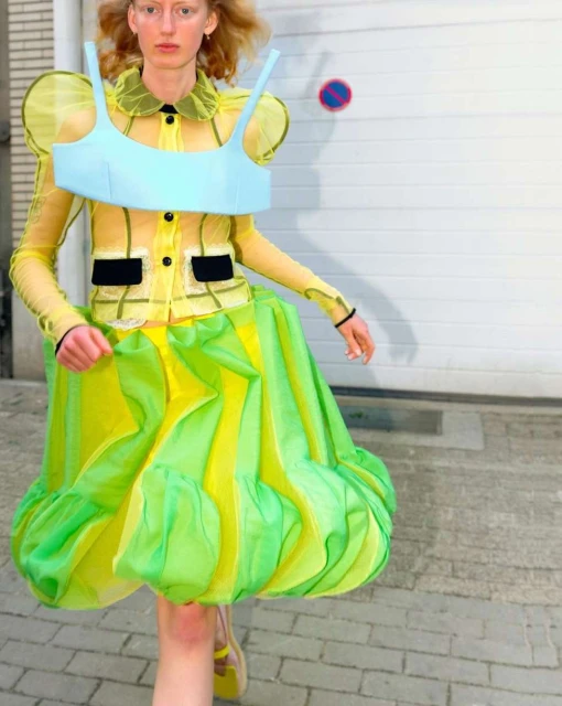Fashion model in a translucent yellow top, blue bra, and balloon skirt posing in an outdoor setting with a garage door background.