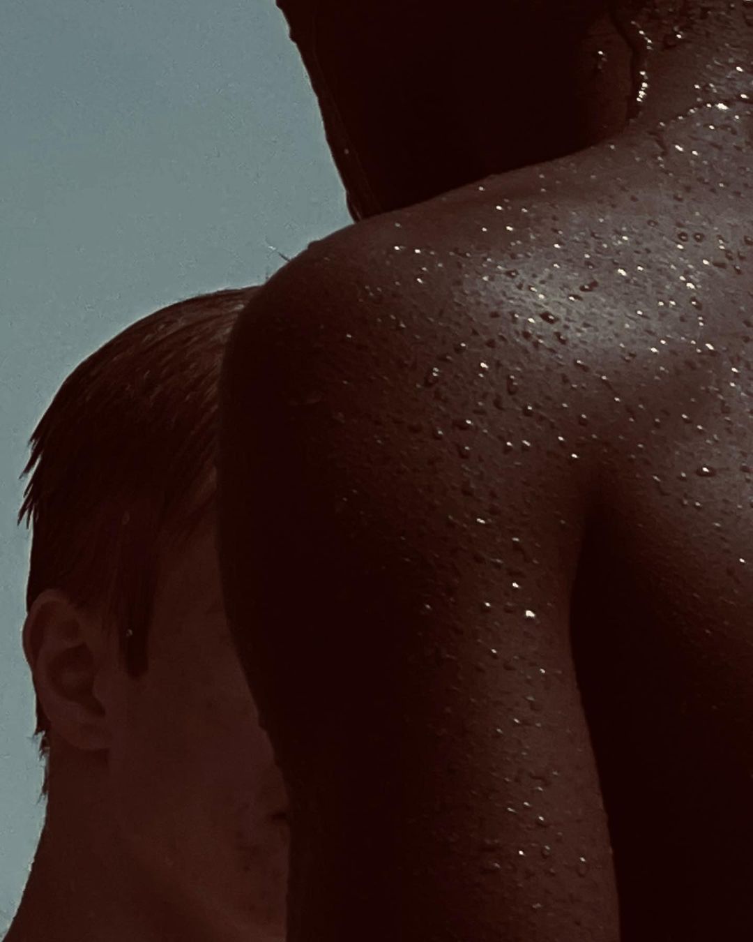 Close-up portrait of two individuals with water droplets on skin against a muted sky background by Mariya Pepelanova
