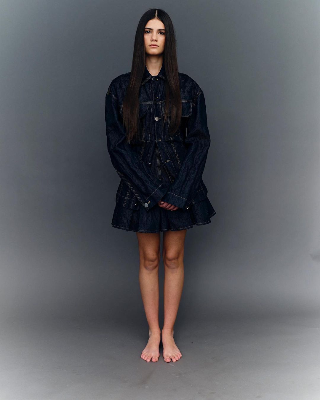 Model with long dark hair wearing a dark denim jacket and skirt set against a grey seamless background