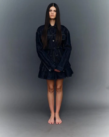 Model with long dark hair wearing a dark denim jacket and skirt set against a grey seamless background