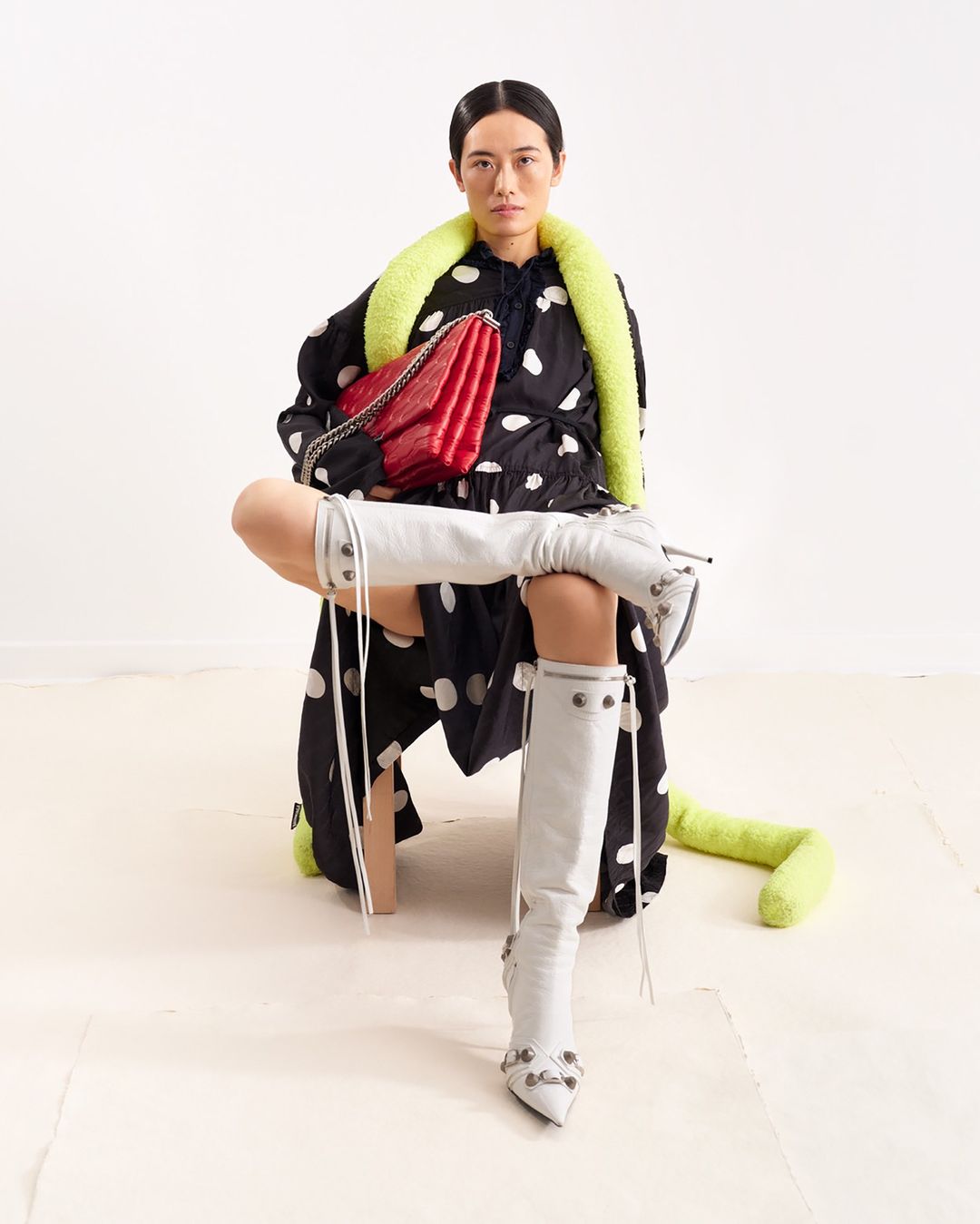 A fashion shot presents a stylish woman in a polka dot dress, knee-high boots, and a red purse, set against a bright, clean backdrop.