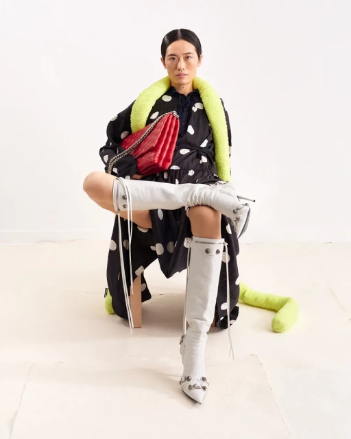 A fashion shot presents a stylish woman in a polka dot dress, knee-high boots, and a red purse, set against a bright, clean backdrop.