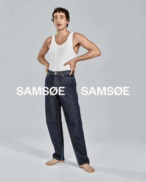 A person poses in jeans and a white tank top in a studio, showcasing a fashion pose with a blurred background, highlighting clothing.