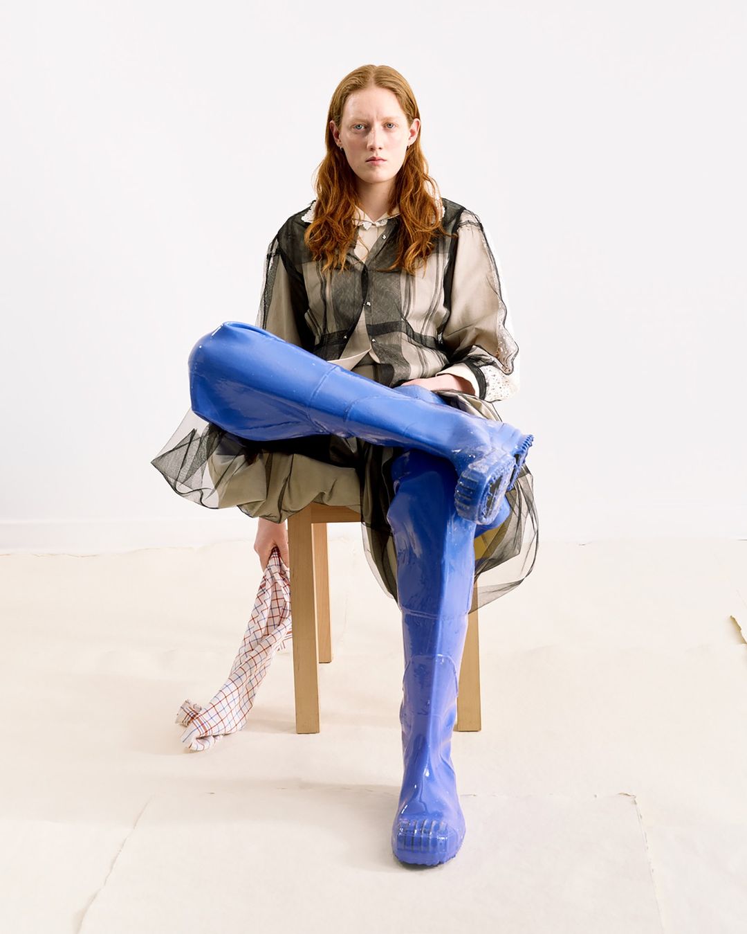 Red-haired woman in transparent shirt dress and bright blue thigh-high boots seated in a minimalist setting, holding checkered cloth.