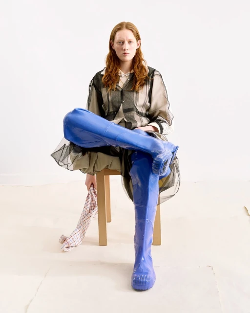 Red-haired woman in transparent shirt dress and bright blue thigh-high boots seated in a minimalist setting, holding checkered cloth.