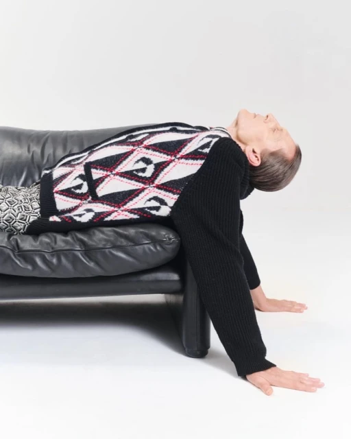 Person in patterned sweater reclining on a black couch in a bright white studio setting with their head tilted back.