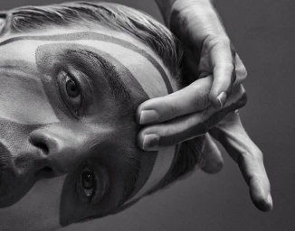 Close-up monochrome portrait of a person with geometric face paint, resting fingers gently on their forehead