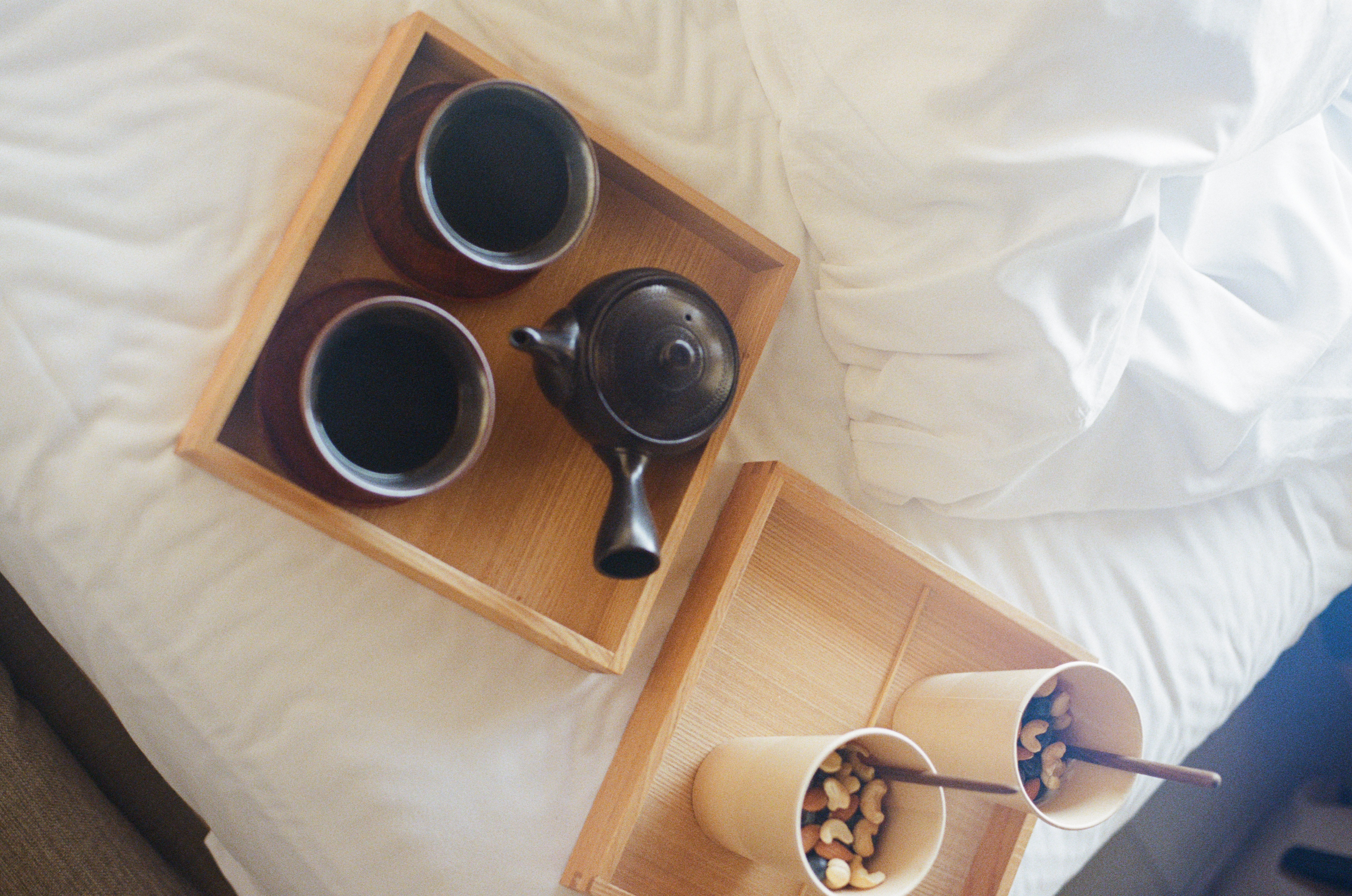 Overhead view of two wooden trays with a teapot, two cups of dark tea, and two cups of nuts on a white bed.
