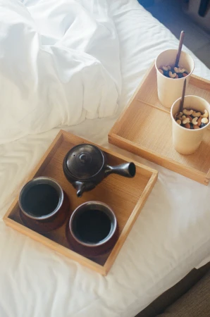 Overhead view of two wooden trays with a teapot, two cups of dark tea, and two cups of nuts on a white bed.