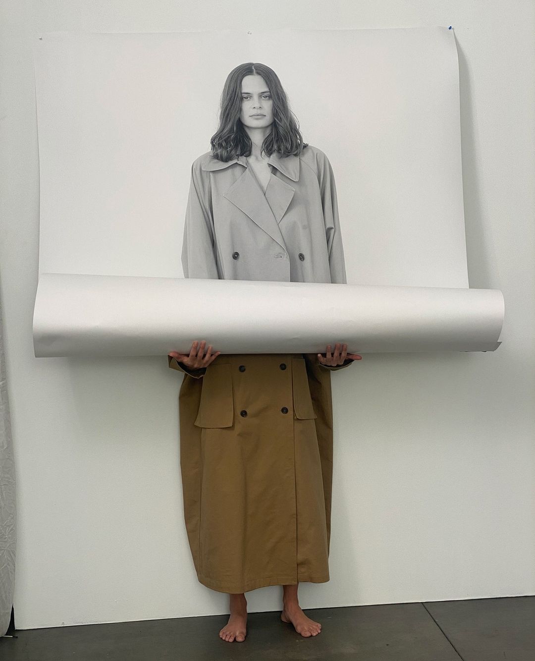 Fashion shoot with a model wearing a trench coat holding a rolled-up poster of a woman, in a minimal photo studio setting.