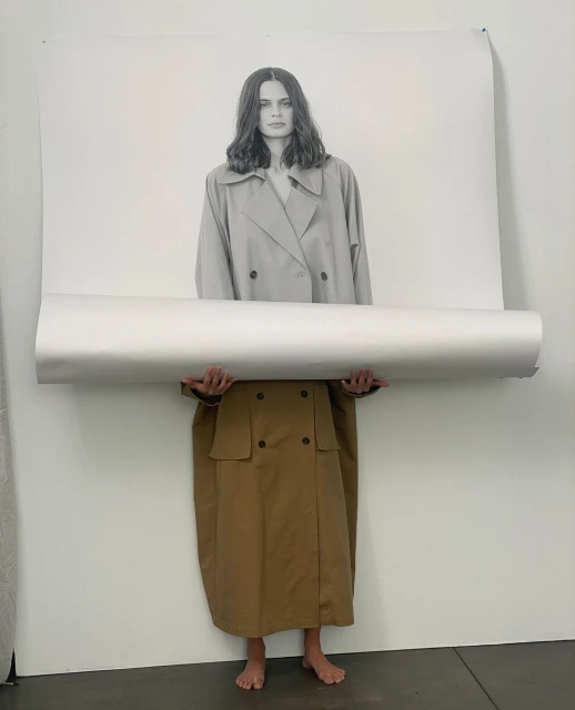 Fashion shoot with a model wearing a trench coat holding a rolled-up poster of a woman, in a minimal photo studio setting.