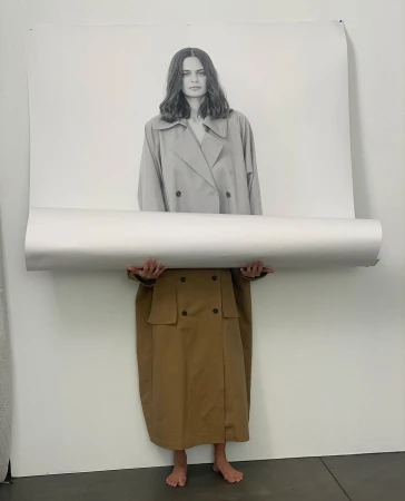 Fashion shoot with a model wearing a trench coat holding a rolled-up poster of a woman, in a minimal photo studio setting.