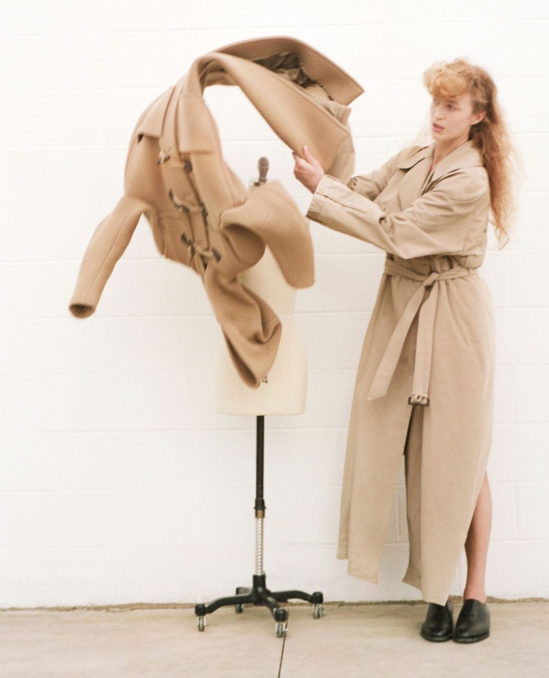 Woman posing in a trench coat standing next to a mannequin with a flowing coat in a minimalist studio with a white wall.