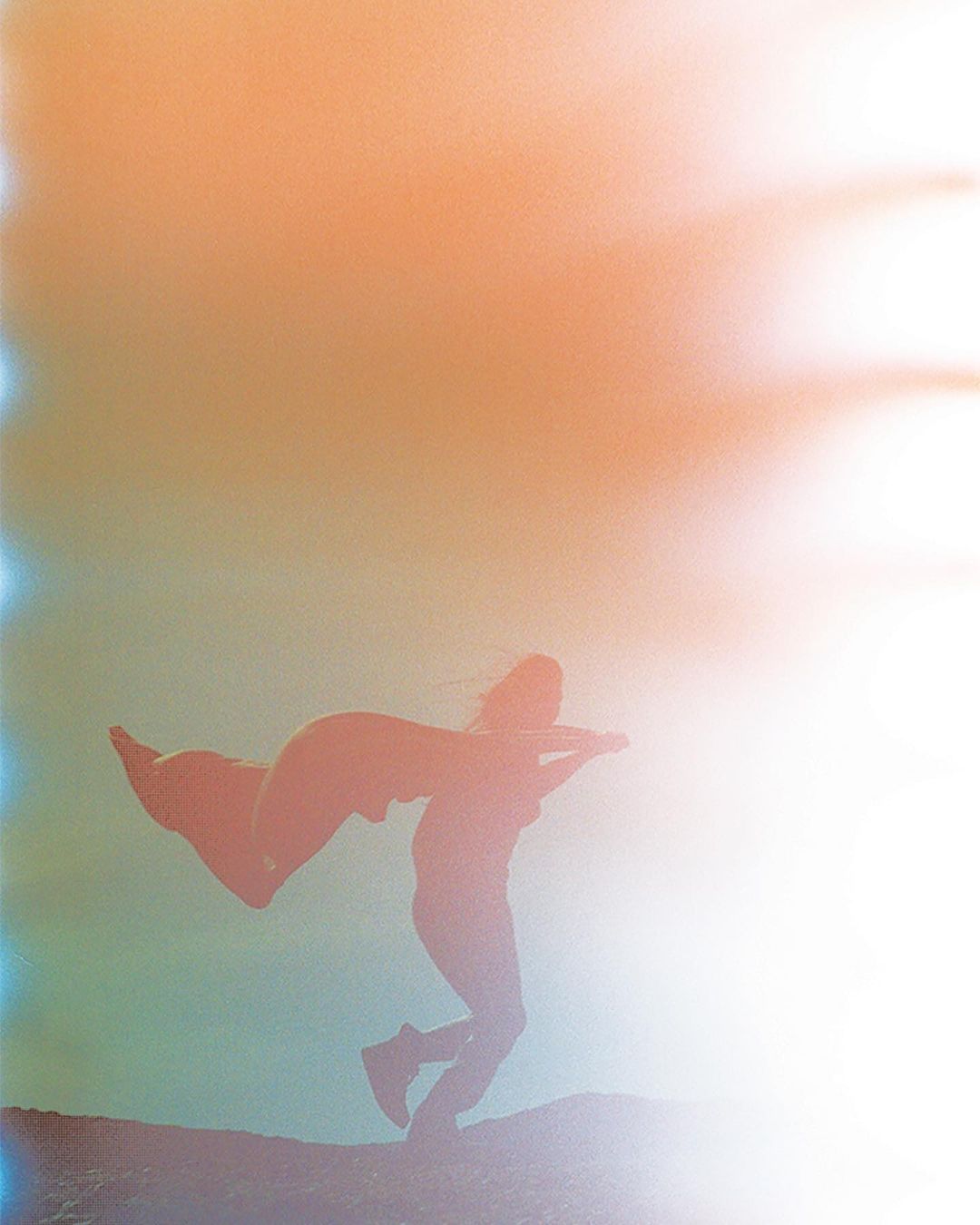 Silhouette of a person wearing a cape jumping in the air on a hill against a vibrant, hazy, peach and blue background.