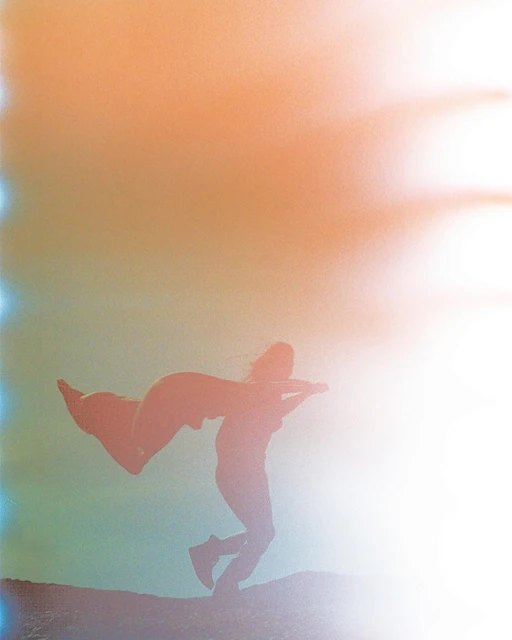 Silhouette of a person wearing a cape jumping in the air on a hill against a vibrant, hazy, peach and blue background.