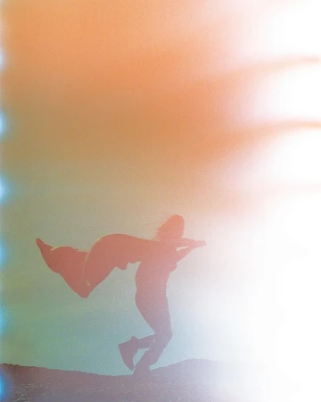 Silhouette of a person wearing a cape jumping in the air on a hill against a vibrant, hazy, peach and blue background.