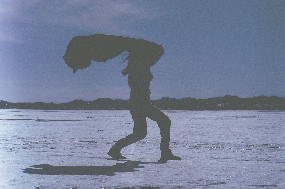 Silhouette of a person standing outdoors on a flat expanse holding a jacket overhead against a pale sky