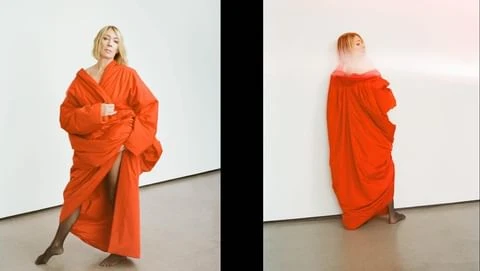 Woman in oversized orange coat poses in a photo studio against a white wall, showcasing fashion and style with a minimalist aesthetic.