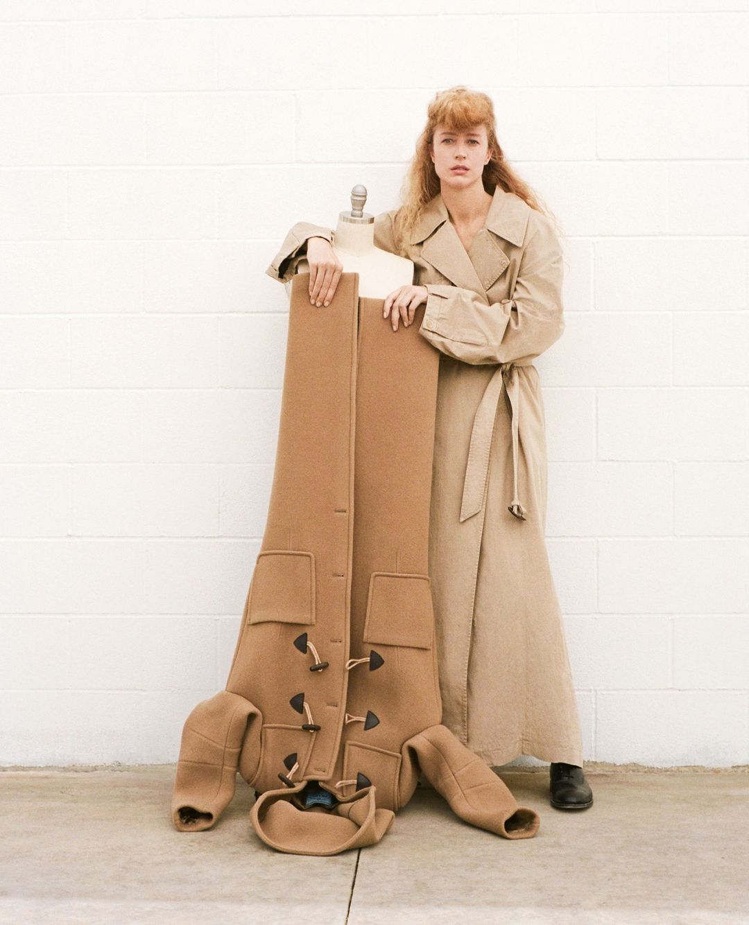 A stylish woman in a trench coat poses with a mannequin displaying a brown coat, against a simple white brick wall and concrete floor.