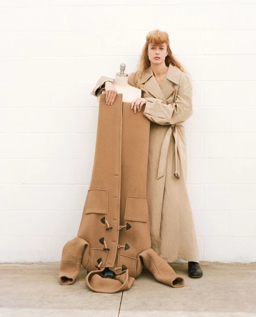 A stylish woman in a trench coat poses with a mannequin displaying a brown coat, against a simple white brick wall and concrete floor.