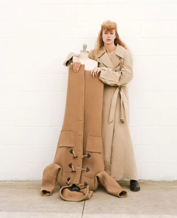 A stylish woman in a trench coat poses with a mannequin displaying a brown coat, against a simple white brick wall and concrete floor.